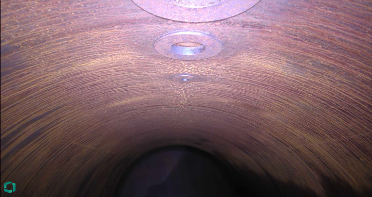 Interior view of a cylindrical freight railcar showing a weathered metallic surface with rust patterns and circular metal fixtures evenly spaced along the inner wall. The lighting accentuates the corroded texture, highlighting the industrial wear typical of bulk transport railcars.