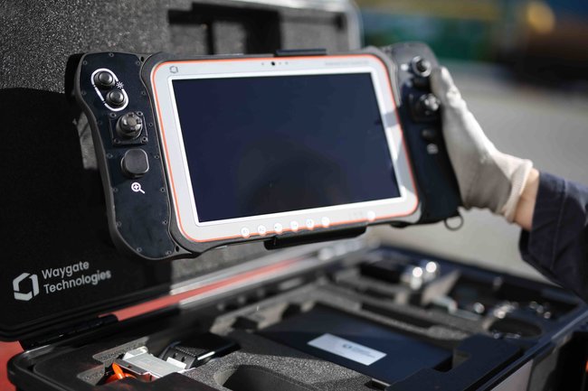 Close up of the Ca-Zoom HD ptz camera operating tablet being held by inspector on a nuclear site. The screen has controls (joy sticks and buttons) on either side and a large display screen in between.