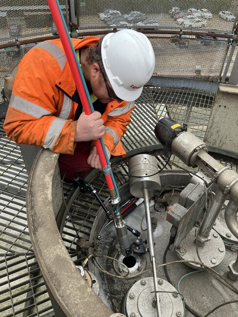 A worker wearing an orange safety jacket and a white hard hat is using a long pole tool to inspect or maintain a brewery tank, likely a fermentation tank. The tool is inserted into an opening in the tank. The setting appears to be a brewery facility with metal grating and various mechanical components visible.