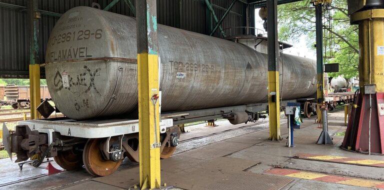 A large cylindrical rail car tank labeled "FLAMAVEL" and numbered "296129-6" is positioned on railway tracks inside a maintenance facility. The tank is elevated on yellow support pillars beneath a metal roof with green structural beams. Various maintenance tools and equipment are visible around the area, indicating an active inspection or repair environment.