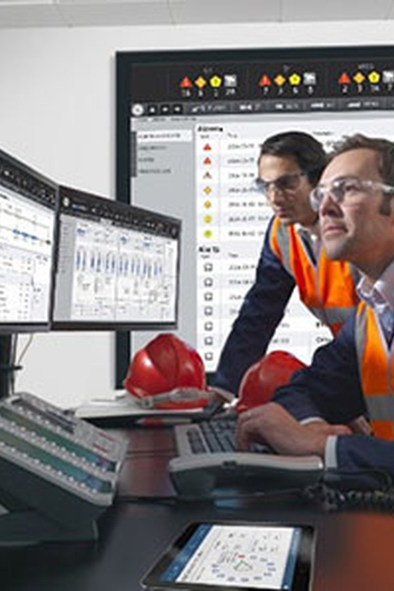 Two workers safety vests and protective glasses looking at multiple screens