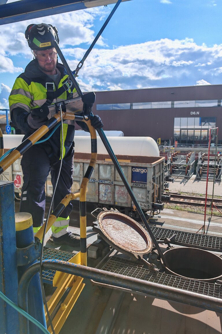 Industrial rail yard scene featuring a worker in reflective safety gear and hard hat managing hose connections atop a freight railcar. The setting includes multiple train cars, a large warehouse structure, and a clear sky with scattered clouds, emphasizing transportation and logistics operations.